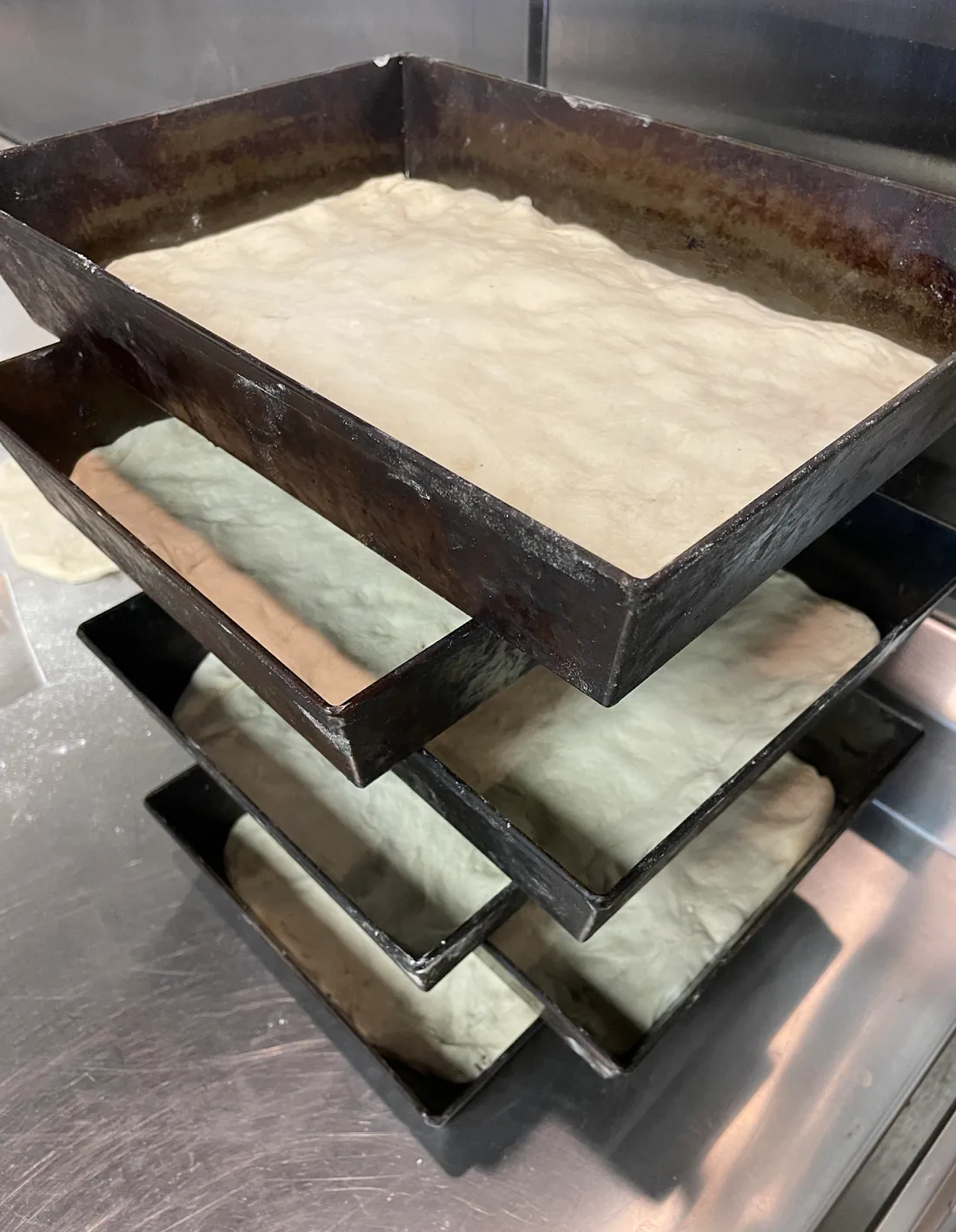 Stacked blue-steel Detroit pizza pans holding dough as it proofs on a stainless prep counter.